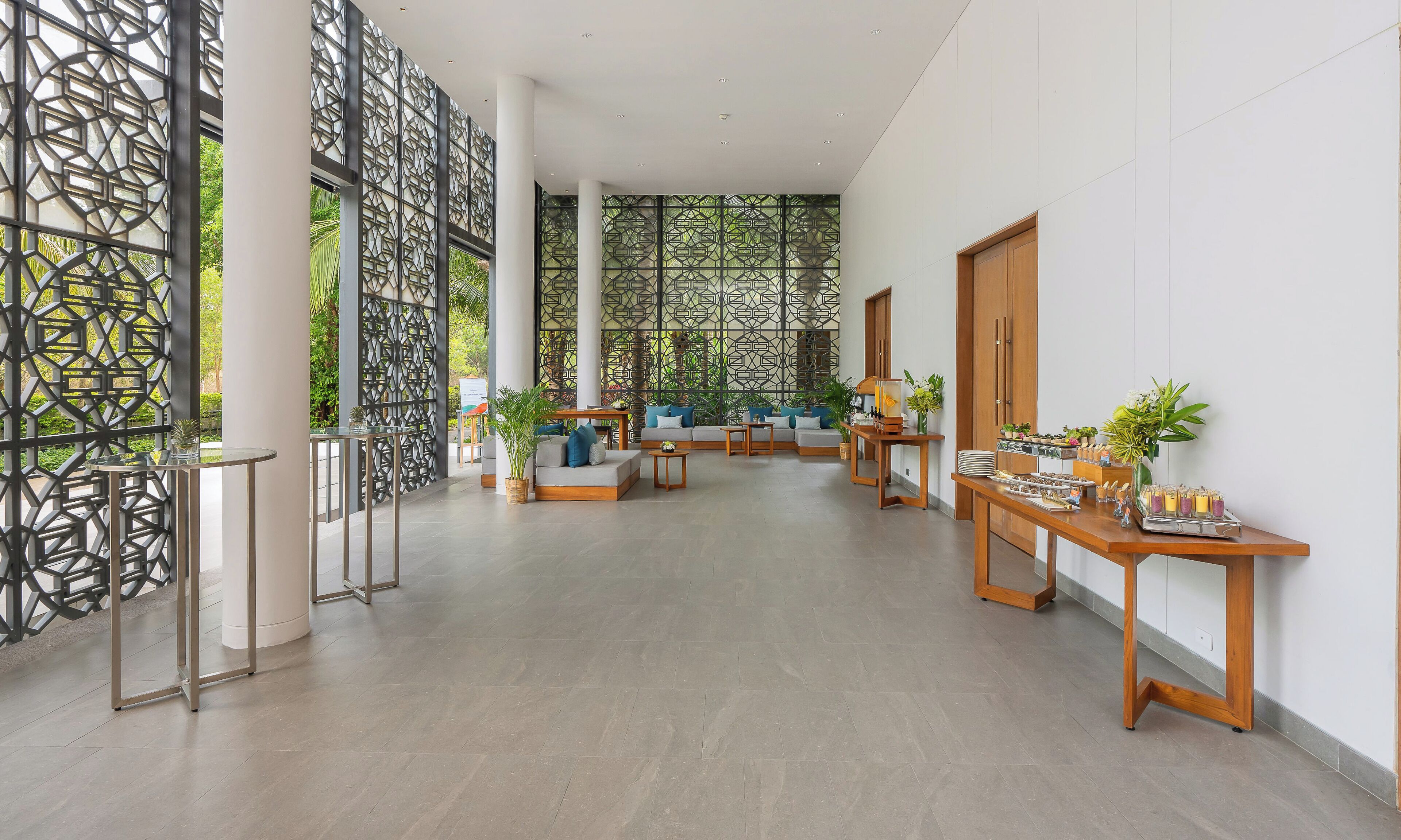 A spacious lobby with a modern design, featuring a seating area with sofas and tables, a decorative wall with geometric patterns, and a view of greenery outside.