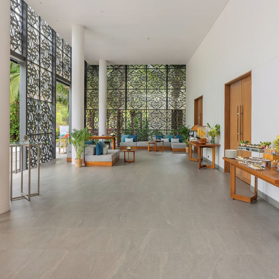 A spacious lobby with a modern design, featuring a seating area with sofas and tables, a decorative wall with geometric patterns, and a view of greenery outside.