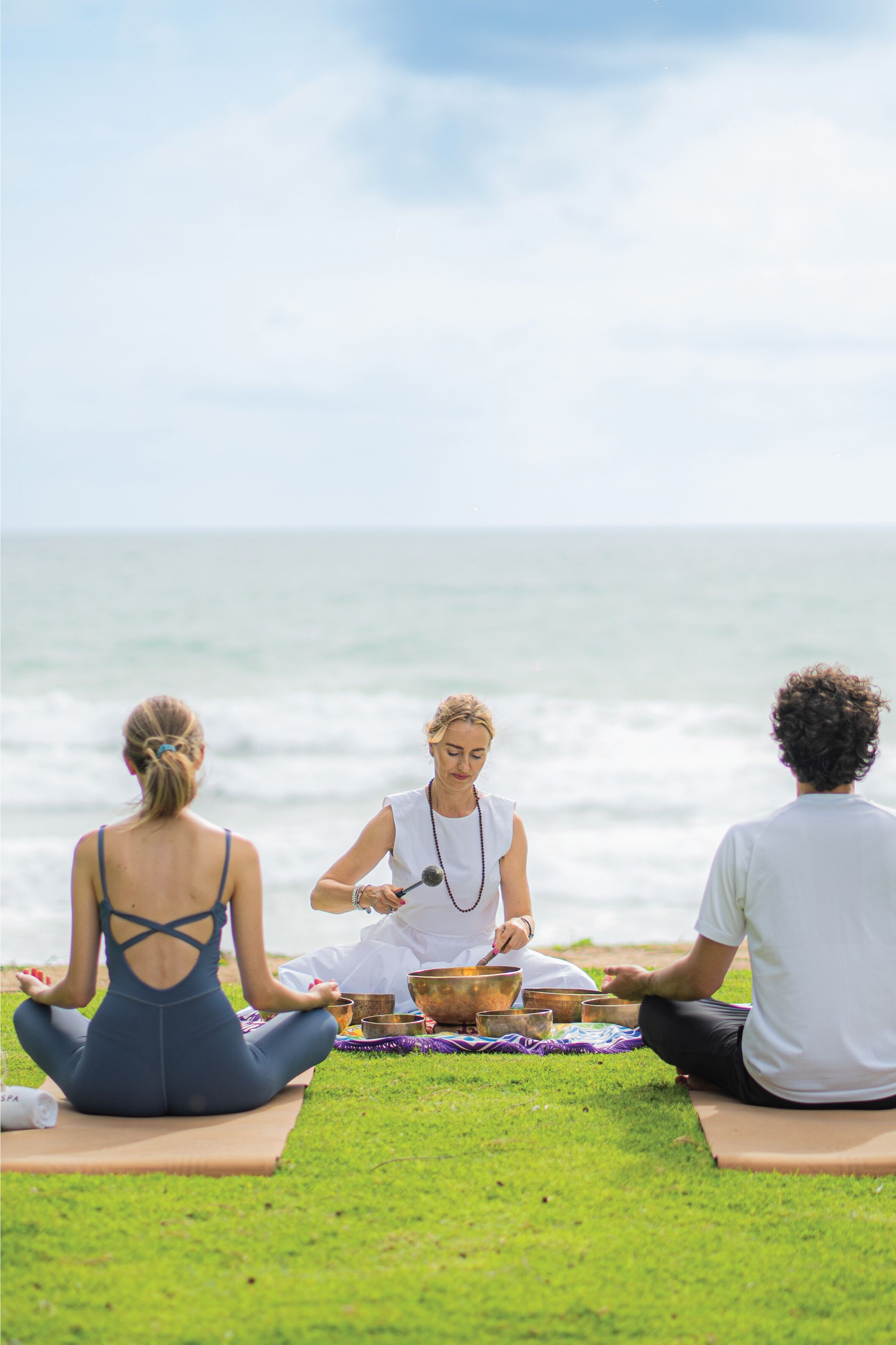 Tres personas meditando en un césped con vista al océano.