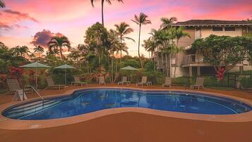 A swimming pool with lounge chairs and umbrellas, surrounded by palm trees and a building with balconies.