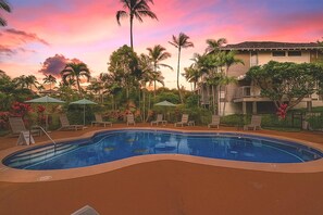 Ein Swimmingpool mit Liegestühlen und Sonnenschirmen, umgeben von Palmen und einem Gebäude mit Balkonen.