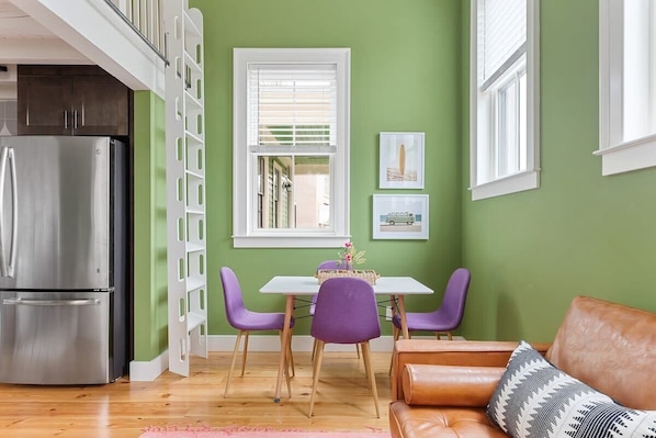 A kitchen with a stainless steel refrigerator, a dining table with purple chairs, and a brown leather sofa.