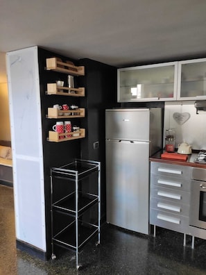 A compact kitchen with a stainless steel refrigerator, wooden shelves with mugs, and a metal rack.