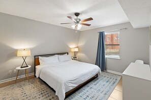 A modern bedroom with a bed, two bedside lamps, a ceiling fan, a window with curtains, and a view of a building outside.