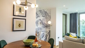 A dining area with a wooden table, green chairs, a green bowl, a bottle, and two glasses. There is a framed insect picture on the wall and a mountain landscape painting.