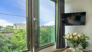 A room with a large window overlooking a historic castle, a flat-screen TV mounted on the wall, and a vase with flowers on a wooden table.