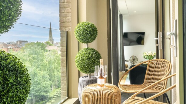 A balcony with a wicker chair, a round table, and two potted plants. A cityscape is visible through the window.