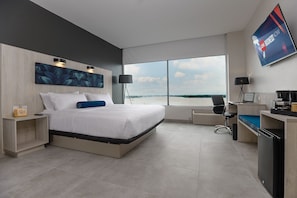 A modern hotel room with a large bed, a desk, a chair, a TV, and a view of the sea. - River Garden Hotel & Suites (Guayaquil)