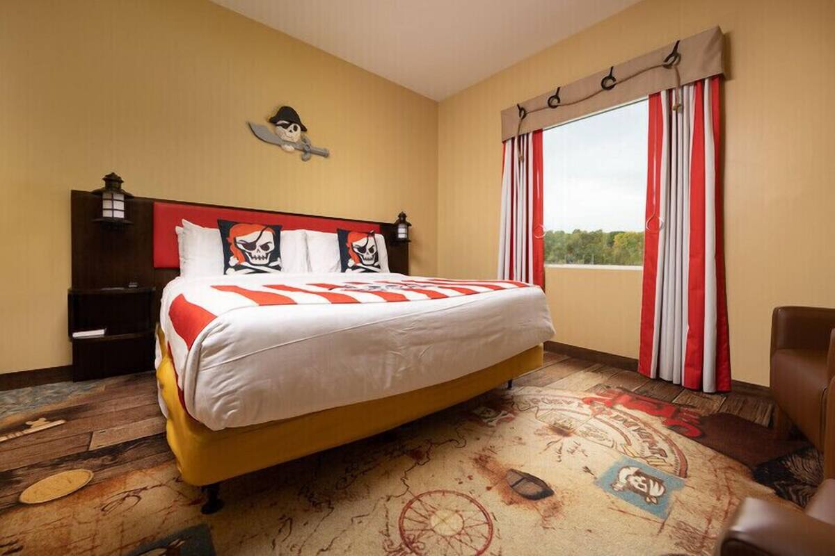 A hotel room with a large bed, a headboard with skull and crossbones decorations, a nightstand, and a window with red and white curtains.