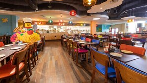 Un restaurante con varias mesas y sillas, una barra central con una figura de cocinero, y luces colgantes de colores.