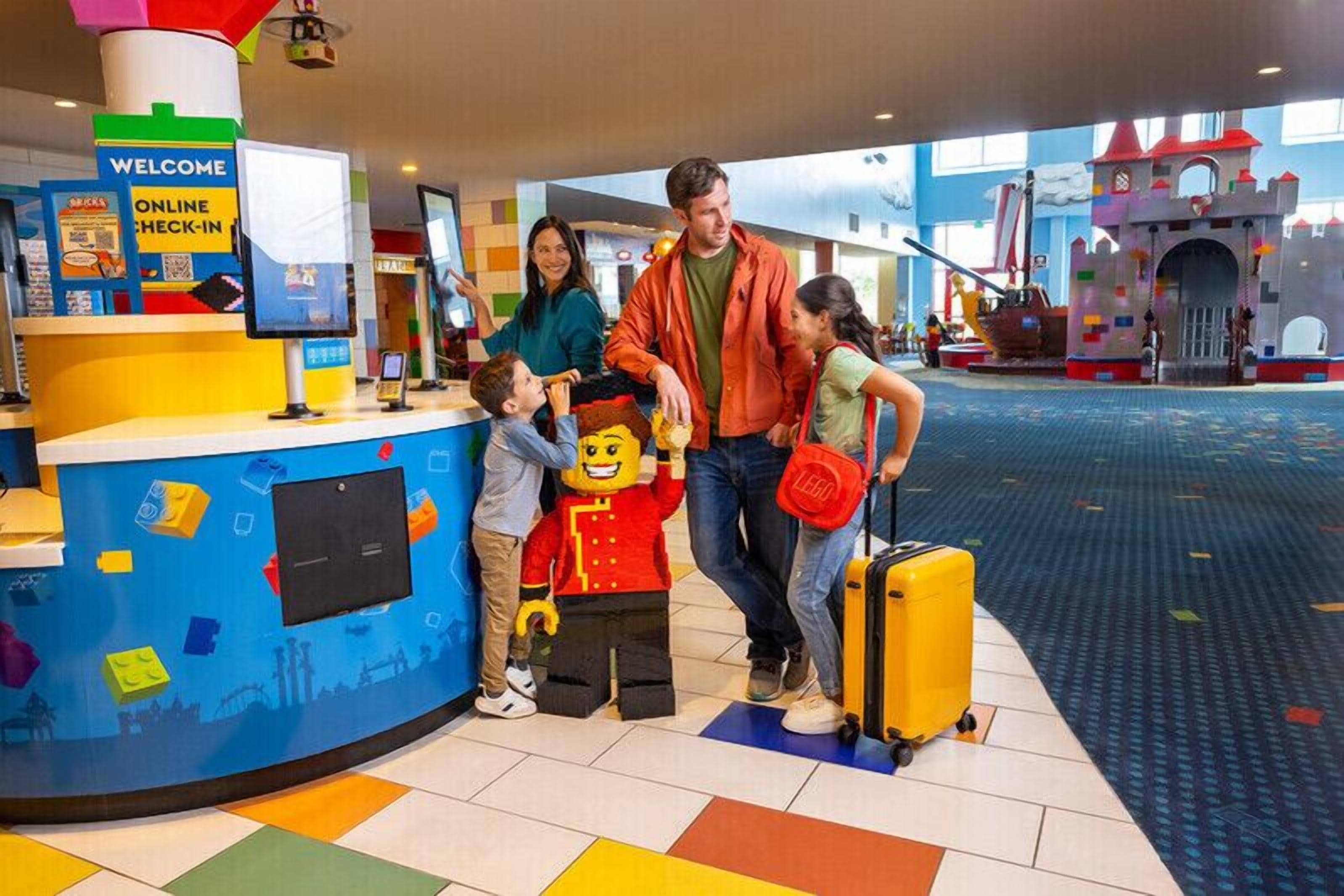 A family interacting with a LEGO-themed kiosk in a brightly lit indoor space with a castle play area in the background.