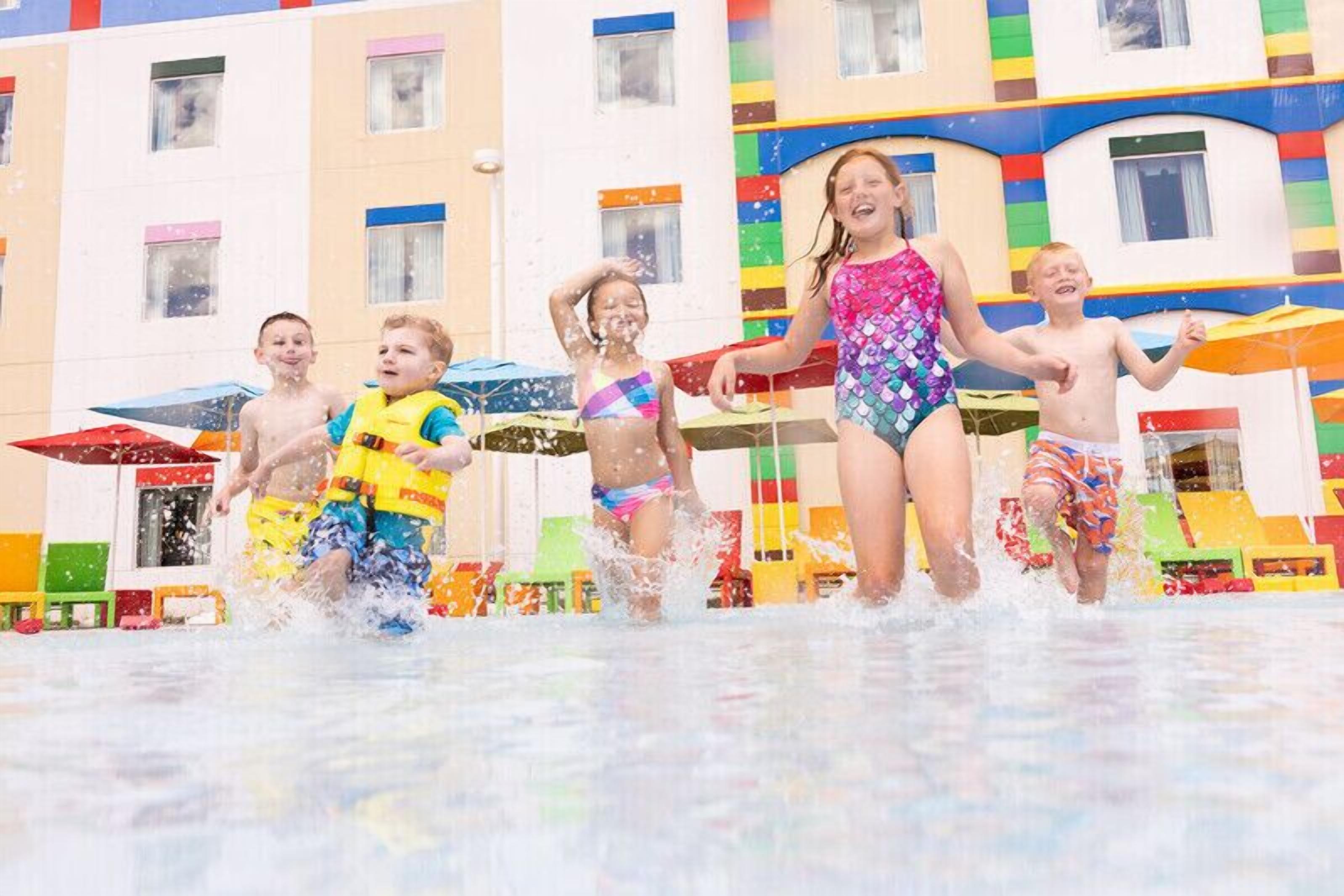 Des enfants jouent dans un parc aquatique, avec des structures de jeux colorées et un bâtiment doté de nombreuses fenêtres en arrière-plan.