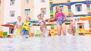 Des enfants jouent dans un parc aquatique, avec des structures de jeux colorées et un bâtiment doté de nombreuses fenêtres en arrière-plan.
