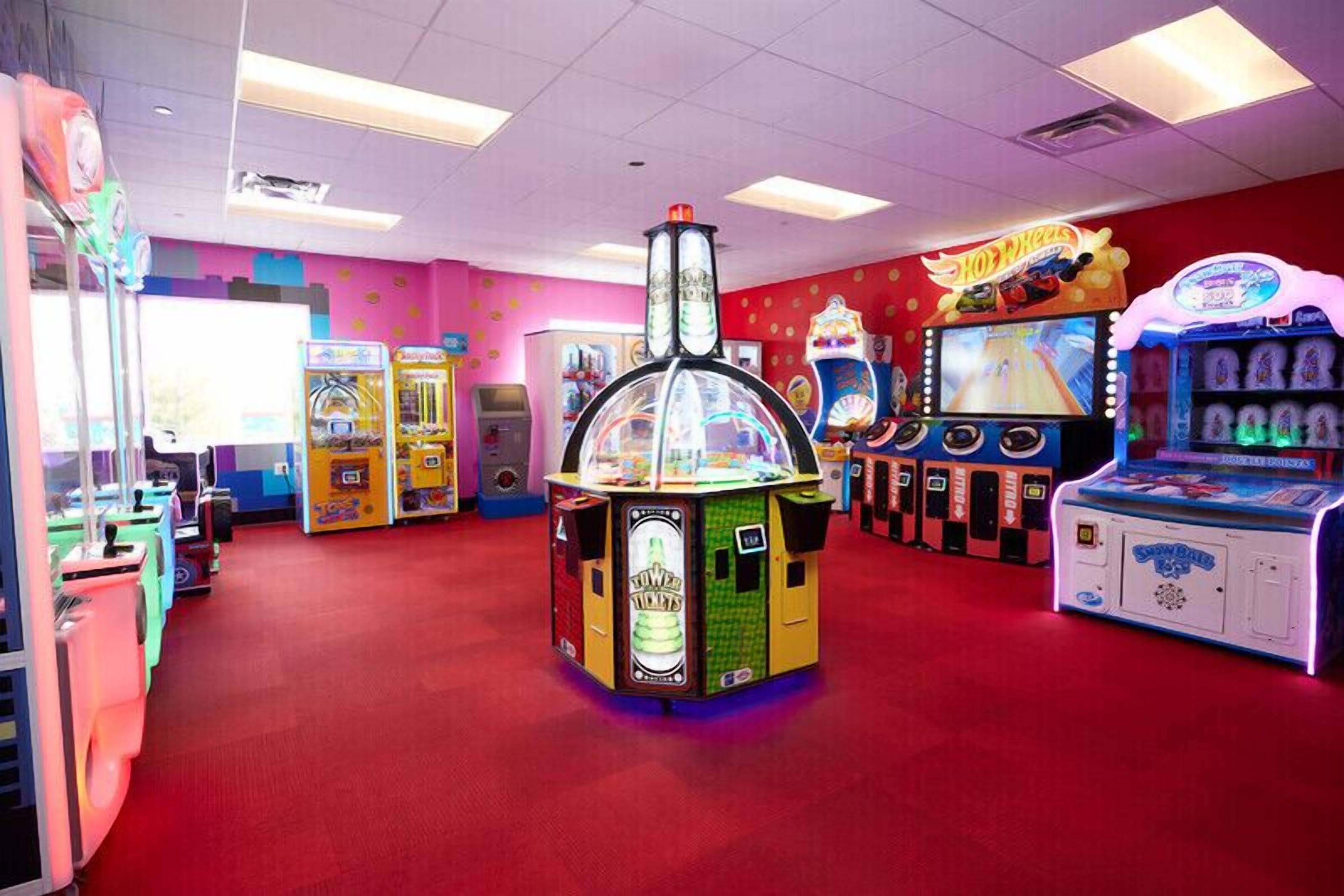 Un salón de juegos bien iluminado con diversas máquinas, incluyendo una máquina destacada con temática 'TOWER' y un juego de 'Hot Wheels'.