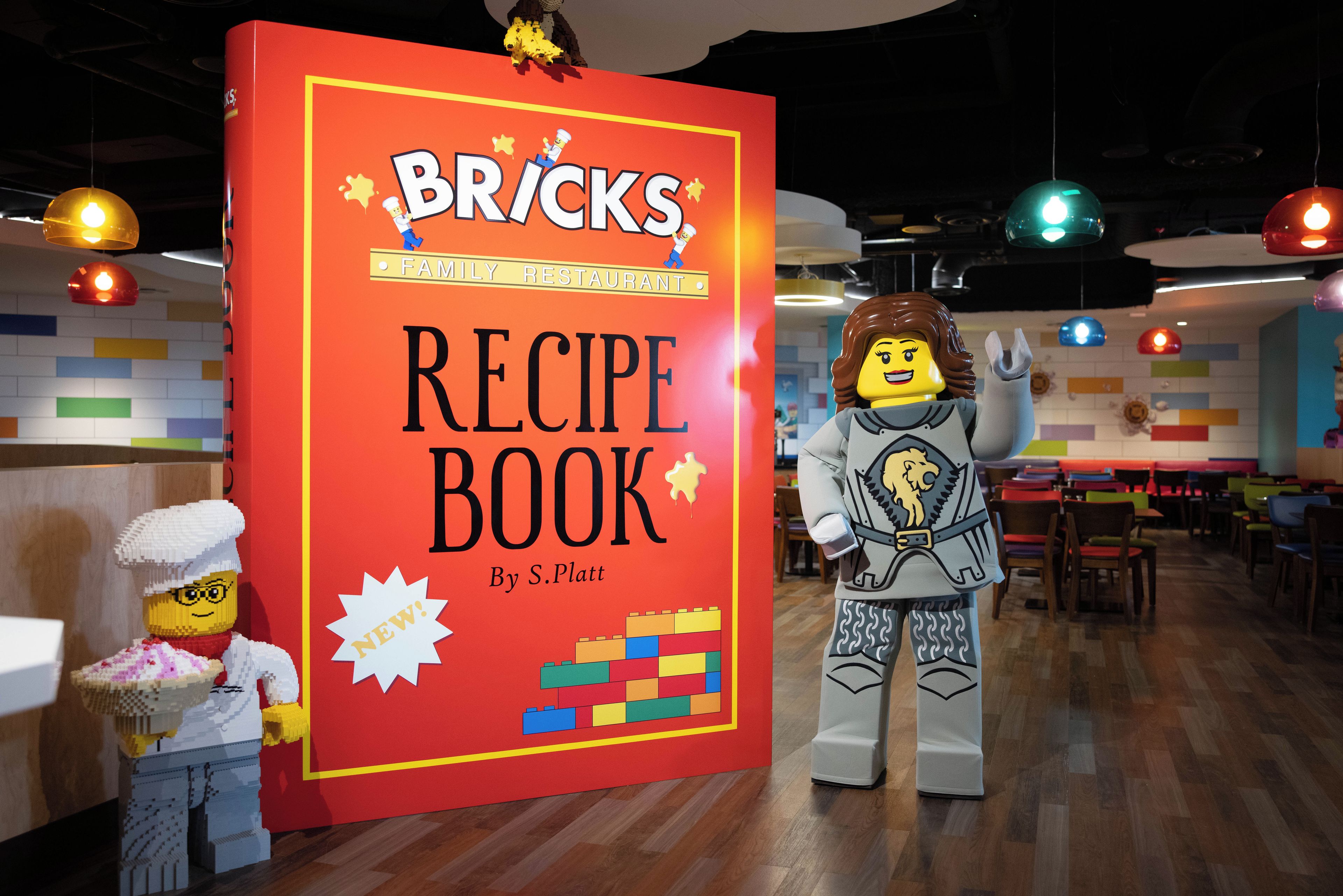A LEGO display with a large red sign reading 'BRICKS RECIPE BOOK' by S. Platt, featuring LEGO figures and a colorful LEGO building.