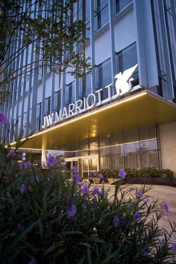 Entrada de um hotel JW Marriott com uma placa chamativa, cercada por flores roxas.