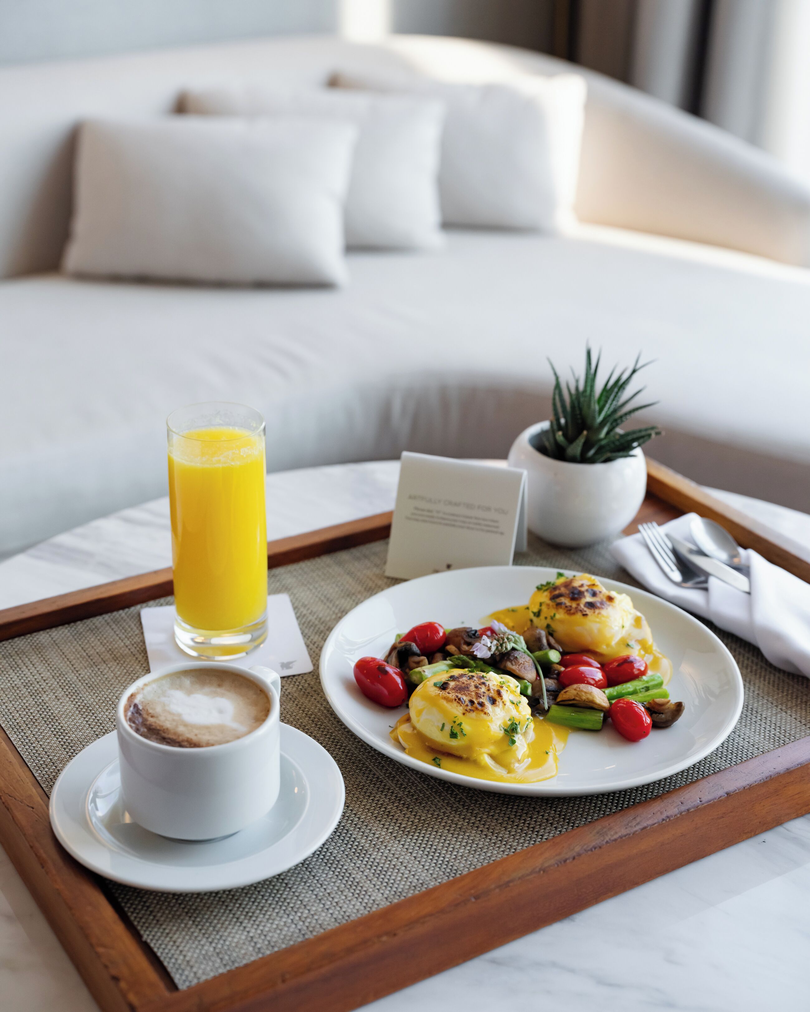 A breakfast tray with eggs benedict, a glass of orange juice, and a cup of coffee on a bed in a hotel room.