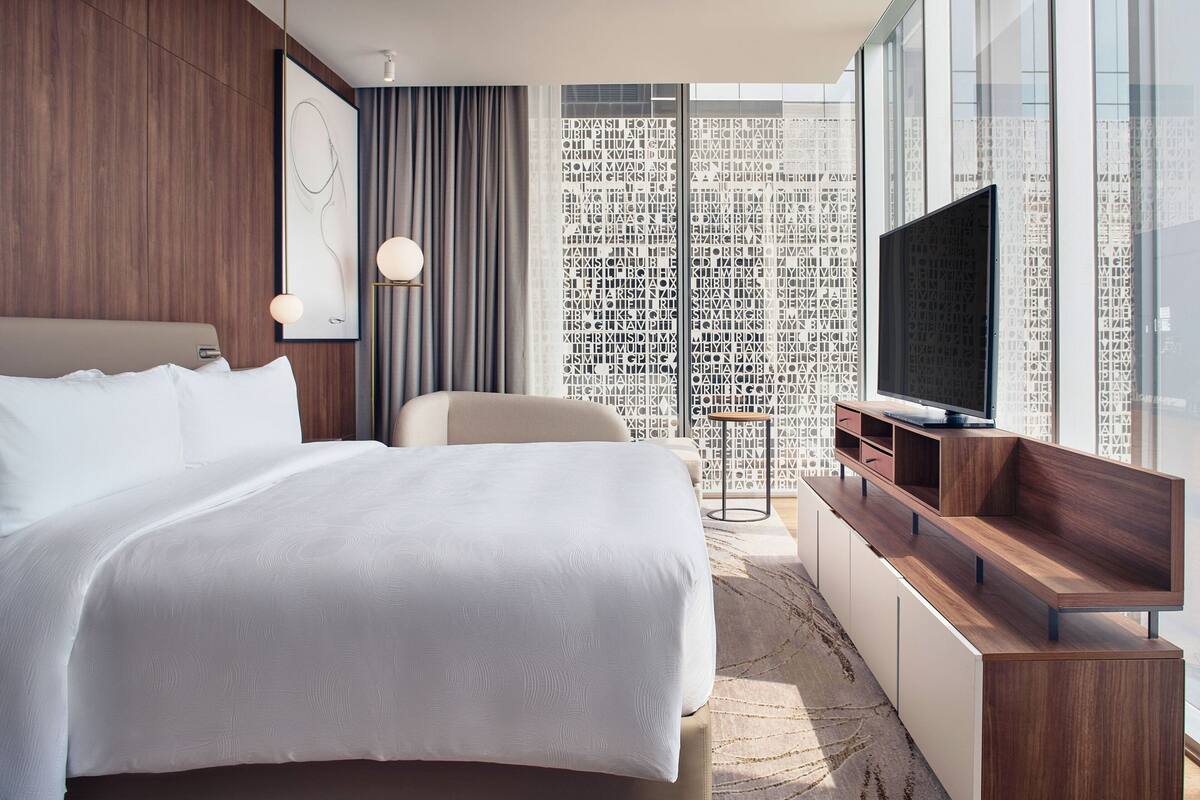Een moderne hotelkamer met een groot bed, een televisie en uitzicht op een stadslandschap door middel van kamerhoge ramen.