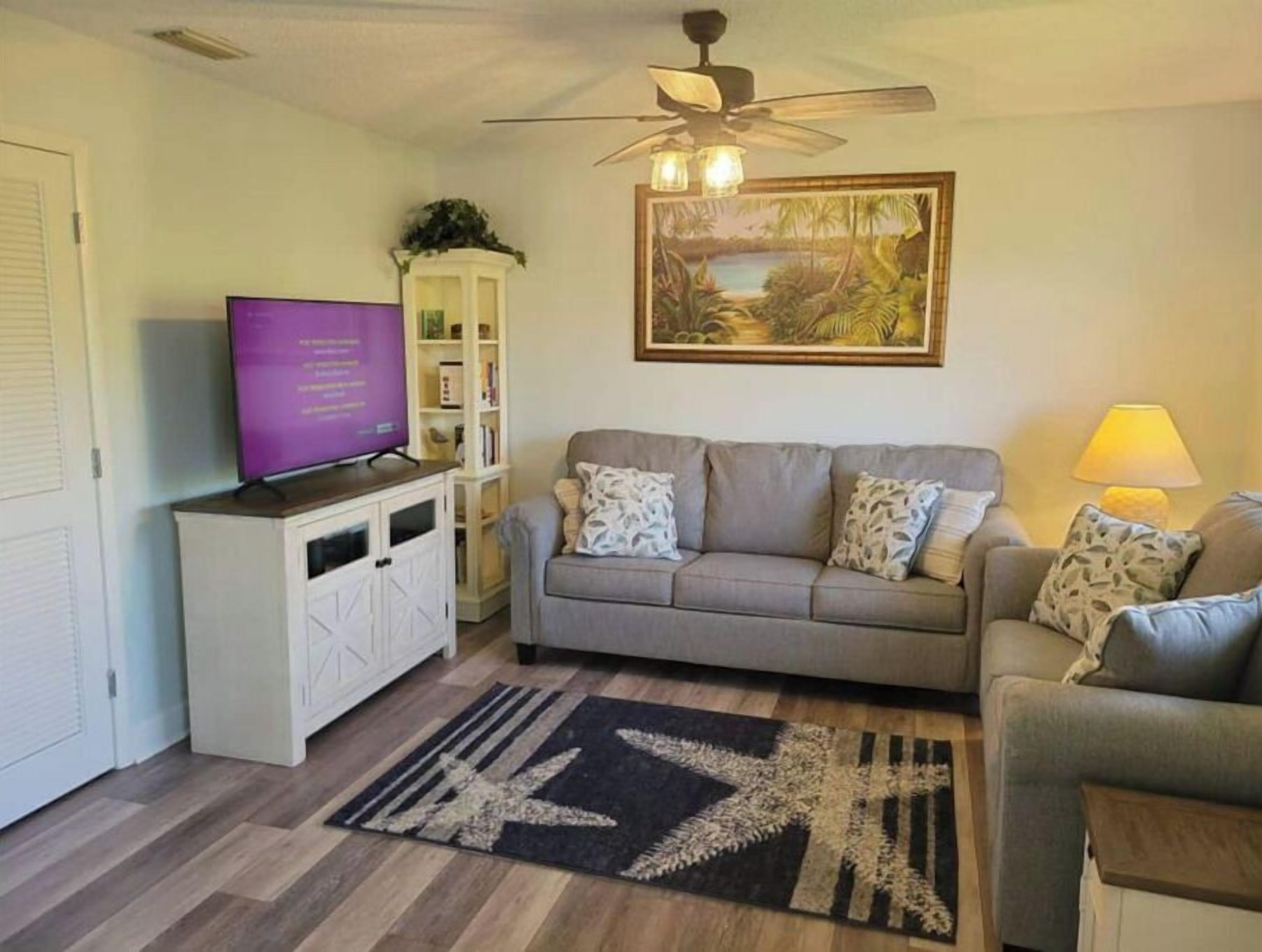 A living room with a grey sofa, a television on a cabinet, a ceiling fan, and a painting of a tropical scene.