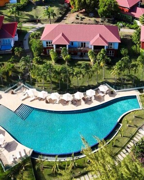 A large, curved swimming pool with lounge chairs and umbrellas, surrounded by palm trees and a building with a red roof. - Tropic Lagoon loft, by the lagoon ! (Le François)
