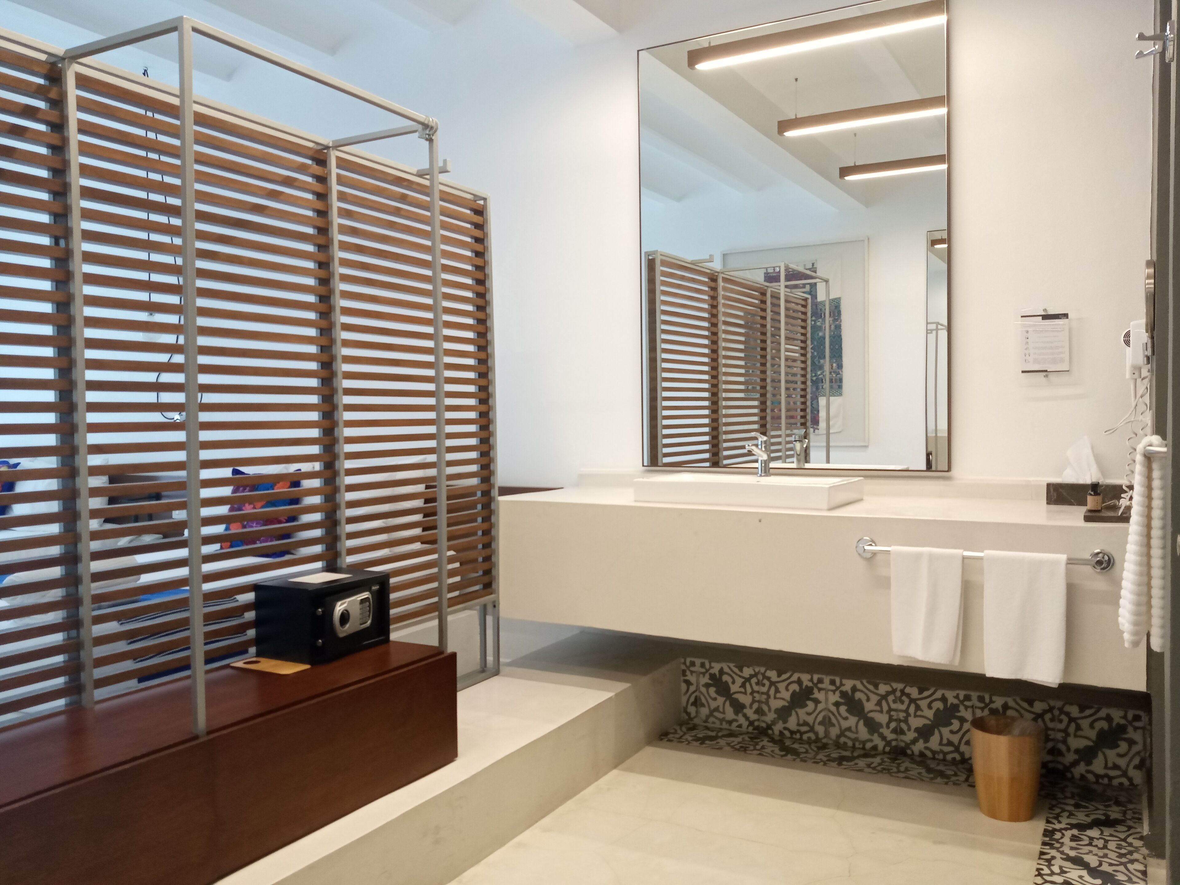 A modern bathroom with a large mirror, wooden slatted blinds, a wooden vanity, and a patterned tile backsplash.