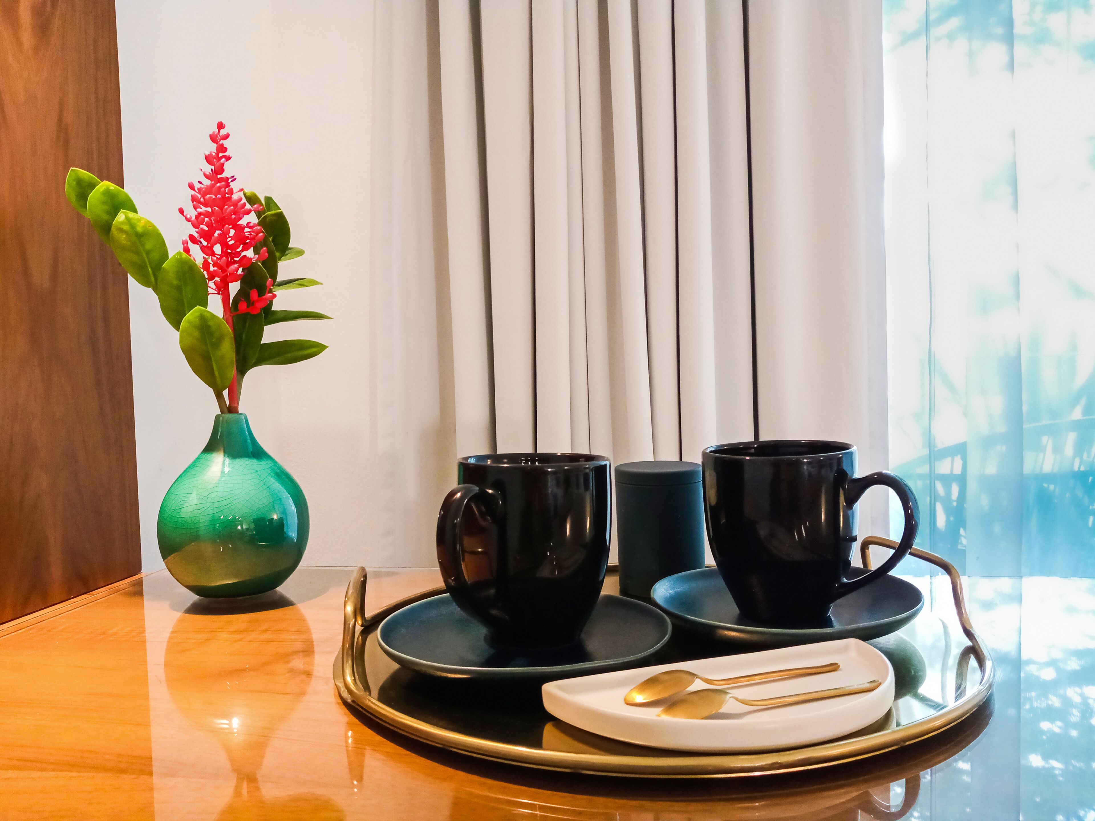 Una mesa con una bandeja que sostiene dos tazas negras, un platillo y una cuchara, y un jarrón verde con flores rojas a la izquierda.