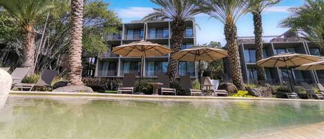 A pool area with lounge chairs and umbrellas, surrounded by palm trees and a building with balconies.