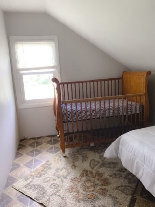 A room with a wooden crib, a bed with a white bedspread, and a patterned rug on a tiled floor.