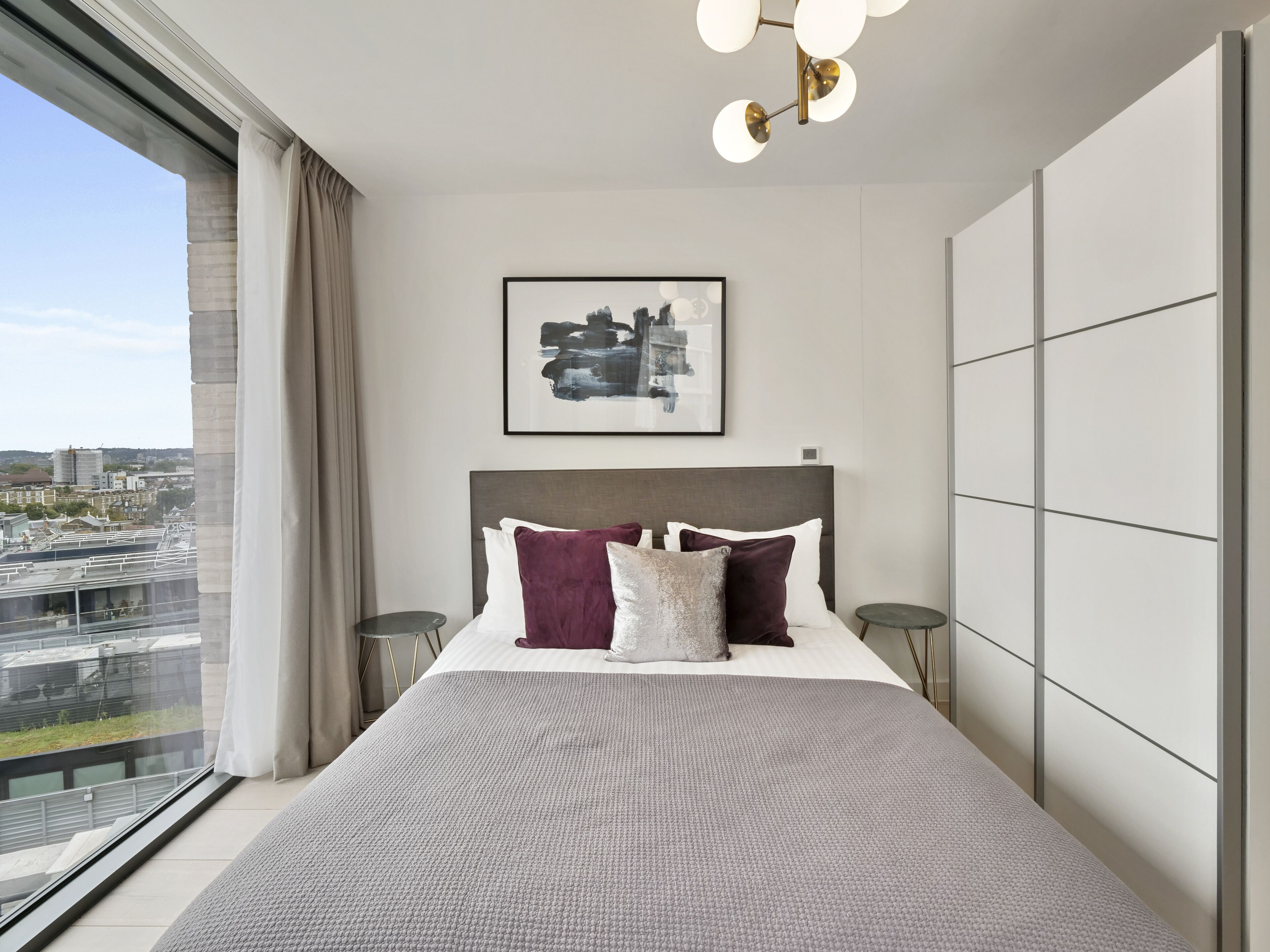 A modern bedroom with a large bed, a city view, and a contemporary light fixture.