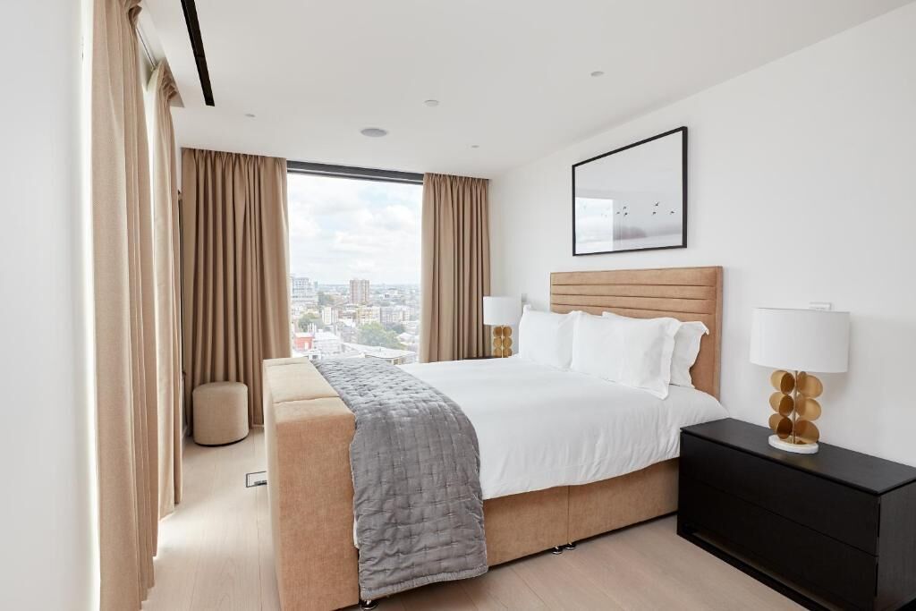 Ein modernes Hotelzimmer mit einem großen Bett, einem Nachttisch, einer Lampe und Blick auf die Stadt durch große Fenster.