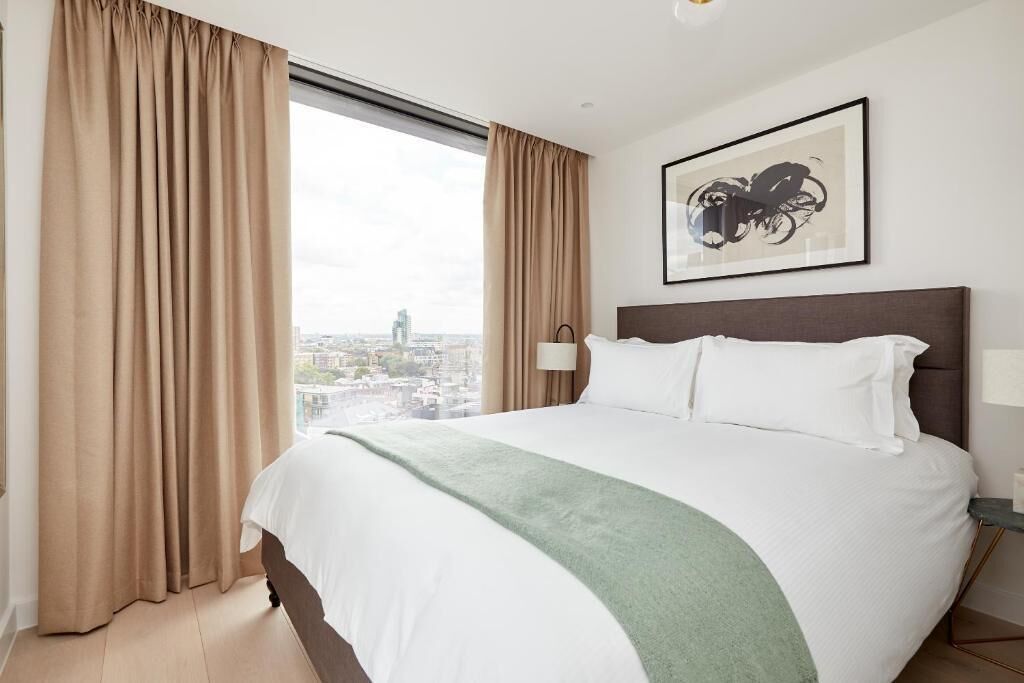 Une chambre d’hôtel avec un grand lit, des rideaux beiges, une vue sur la ville et une œuvre d’art encadrée au mur.