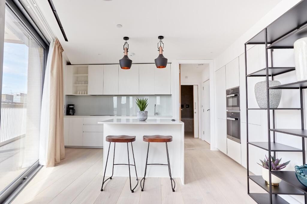 A modern kitchen with white cabinets, a central island with stools, pendant lighting, and a view through a large window.