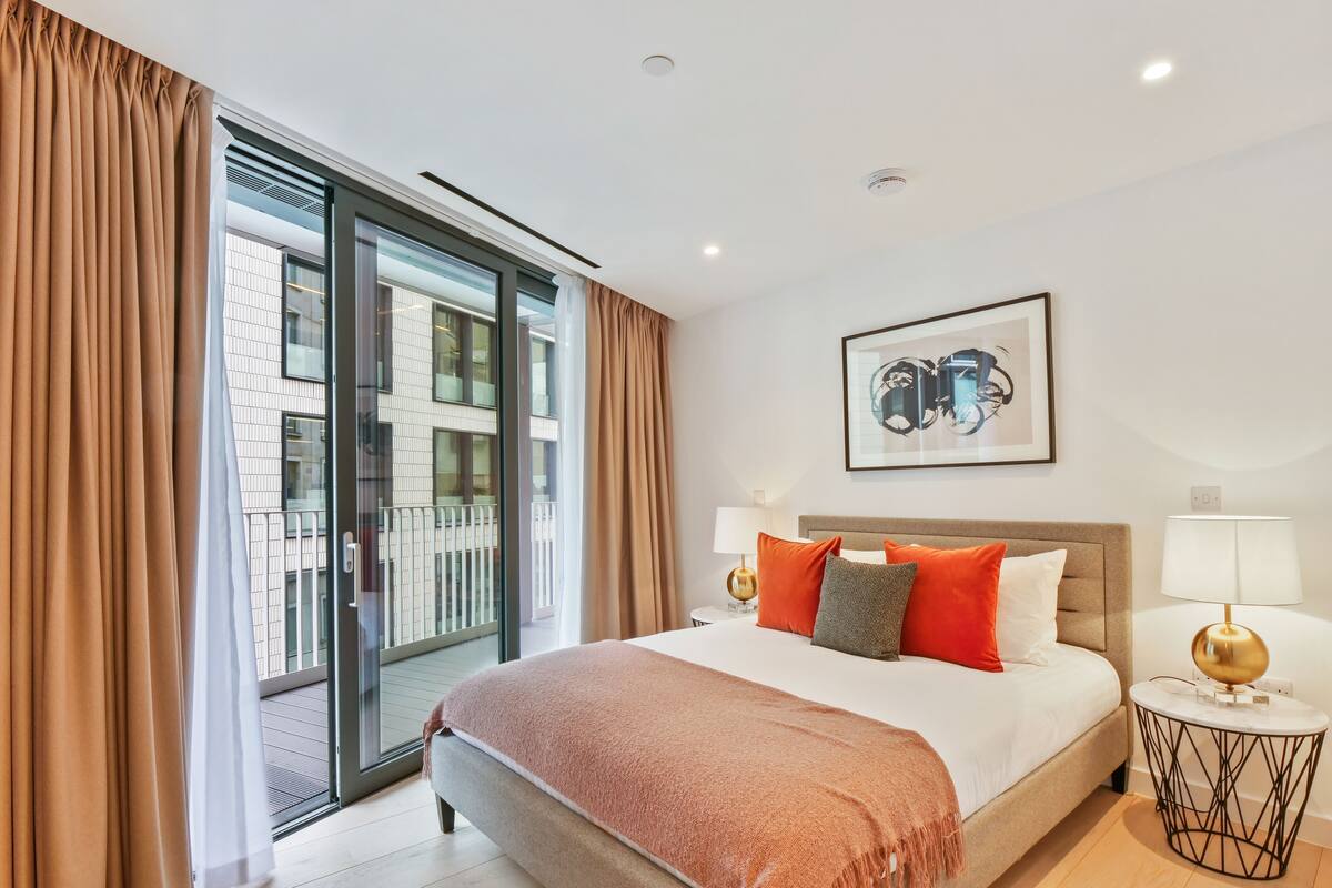 A hotel room with a large bed, a view through a sliding glass door, a framed artwork on the wall, and a round side table with a lamp.