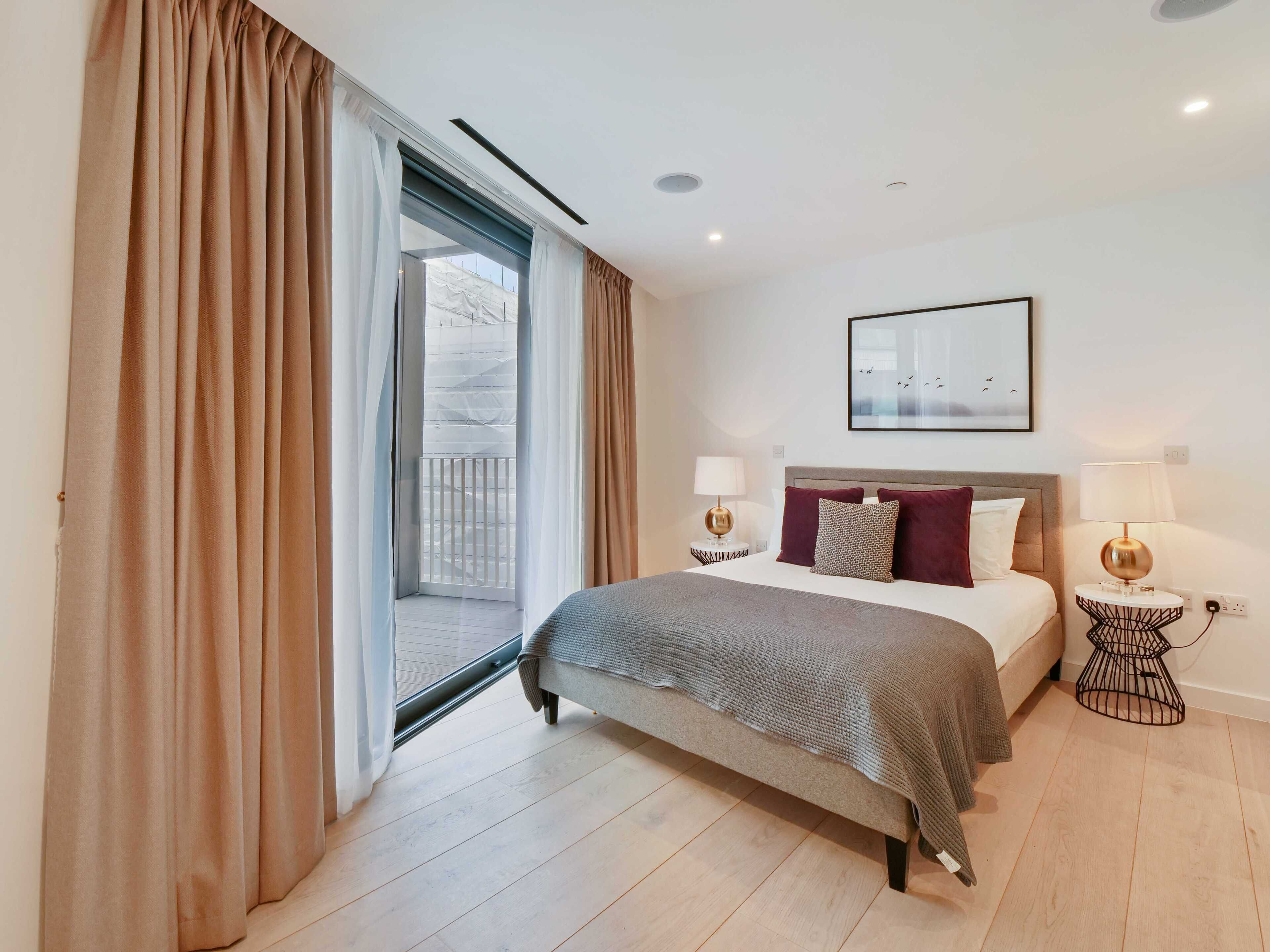 A hotel room with a bed, two bedside tables with lamps, a window with curtains, a framed picture on the wall, and a balcony visible through the window.
