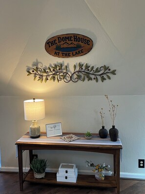 A wooden table with a lamp, a sign reading 'The Dome House at the Lake,' decorative metal leaves, and small potted plants. - Tranquil Lake View Dome Home in Wine Country (Lower Lake)