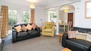 A spacious living room with a black leather sofa, a yellow armchair, and a wooden coffee table. There is a dining area visible through an archway. - Bridgedown House (Richmond)