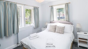 A neatly made bed with white linens, a bedside table with a lamp, and a window with curtains. - Bridgedown House (Richmond)