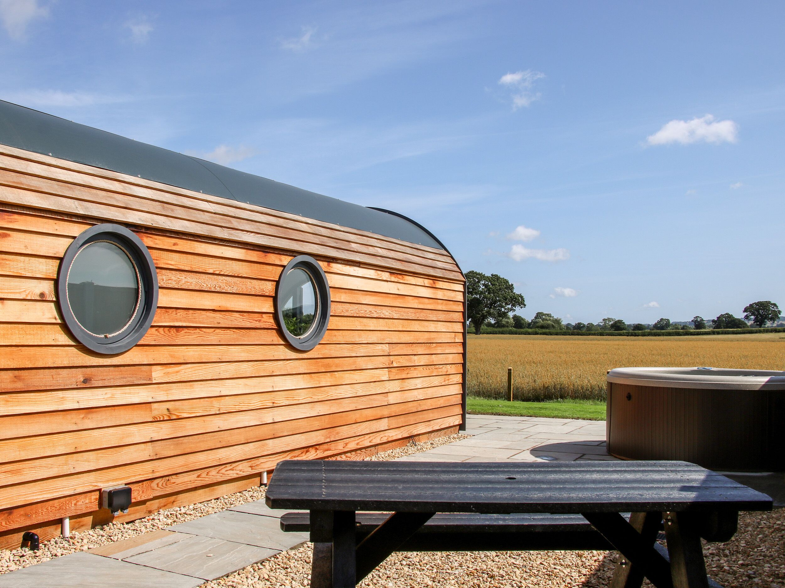 A wooden RV with two round windows, a picnic table, and a hot tub in a rural setting.