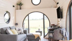 En ombonad inredning med en grå soffa, en liten pall, ett runt bord med en platt-TV och ett stort, välvt fönster som ger utsikt över ett fält.