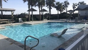 A swimming pool with lounge chairs and umbrellas, surrounded by palm trees and a wooden fence. - 4br/3Ba Oceanview from balcony!  Sleeps 12 w/ hot tub! (Corpus Christi)