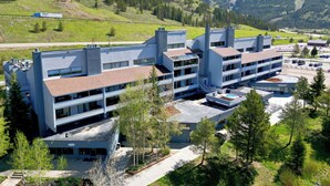 A modern multi-story building with a flat roof, surrounded by greenery and mountains.