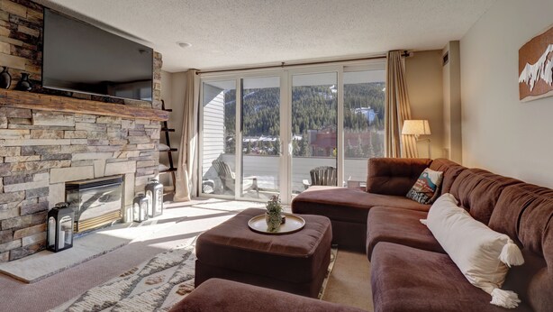 A living room with a stone fireplace, a flat-screen TV, a brown leather sofa, and a large window with a view of a forested area.