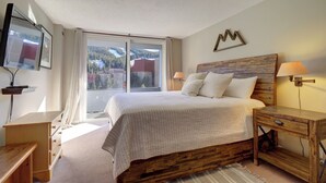 A bedroom with a large bed, wooden headboard, bedside lamps, a TV on a stand, and a view of the outdoors through a window.