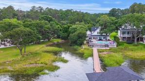 Un muelle a la orilla del río con un área de descanso cubierta, una casa con piscina y un paisaje verde exuberante.