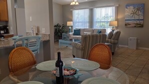 A dining area with a glass table, wicker chairs, and a bottle of wine. There is a kitchen in the background with a refrigerator and a coffee maker.