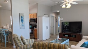 A living room with a sofa, chairs, a glass coffee table, a TV, and a kitchen in the background.