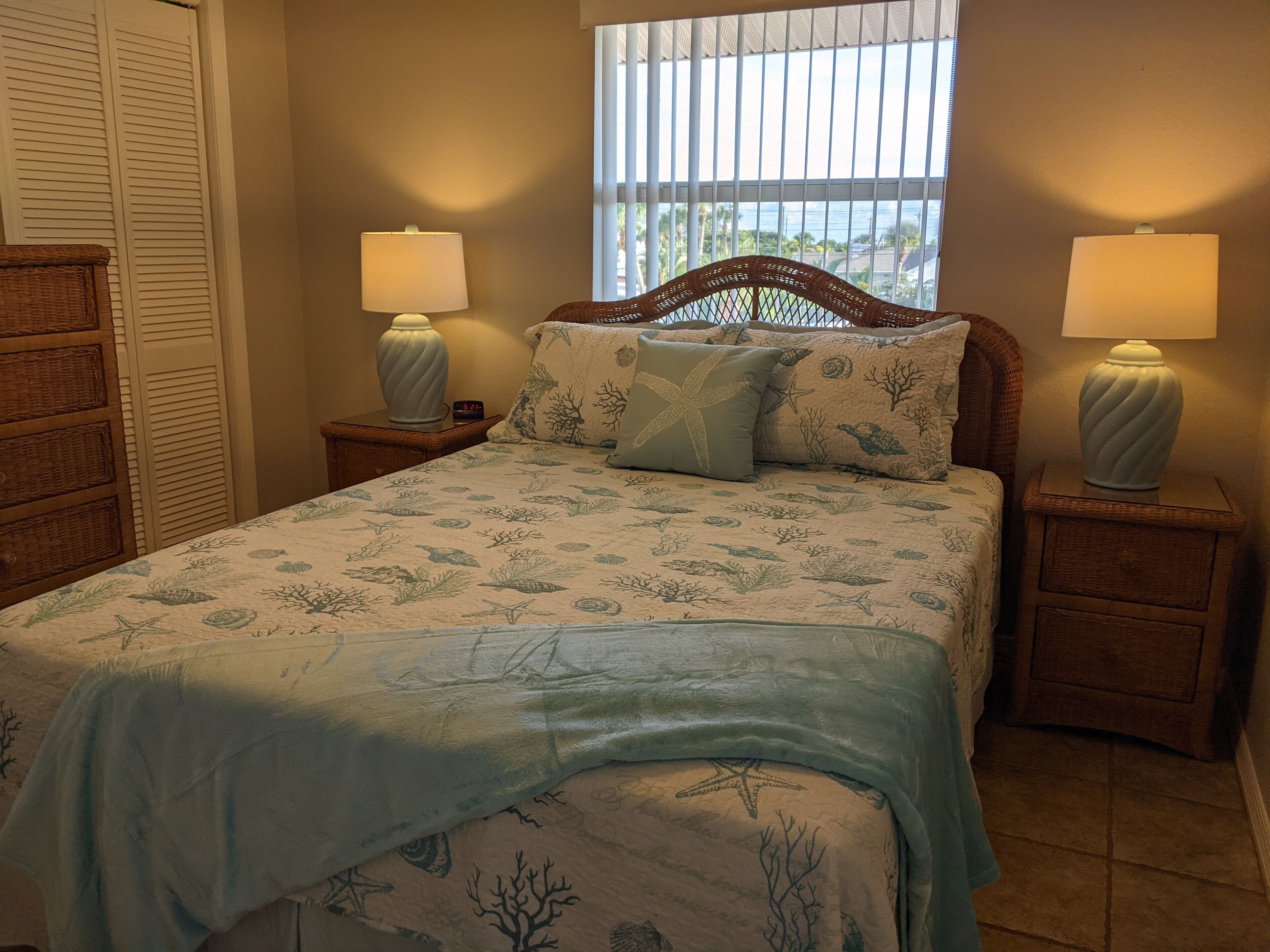A bedroom with a bed, two nightstands, a window with blinds, and a view of the outdoors.