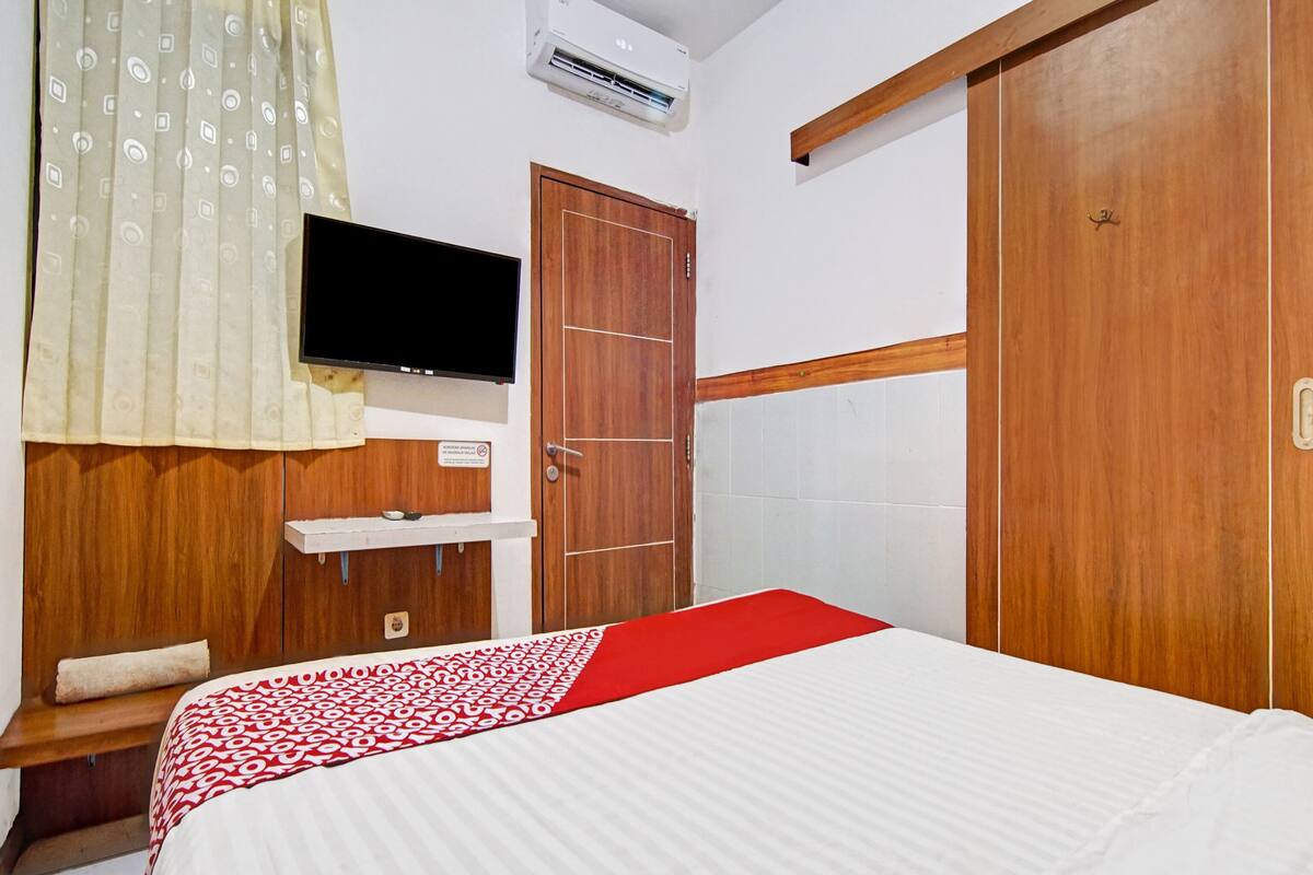 A hotel room with a bed, a TV mounted on the wall, a wooden wardrobe, and a red and white patterned bedspread.