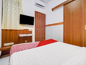 Habitación de hotel con cama, televisor montado en la pared, armario de madera y colcha estampada en rojo y blanco.
