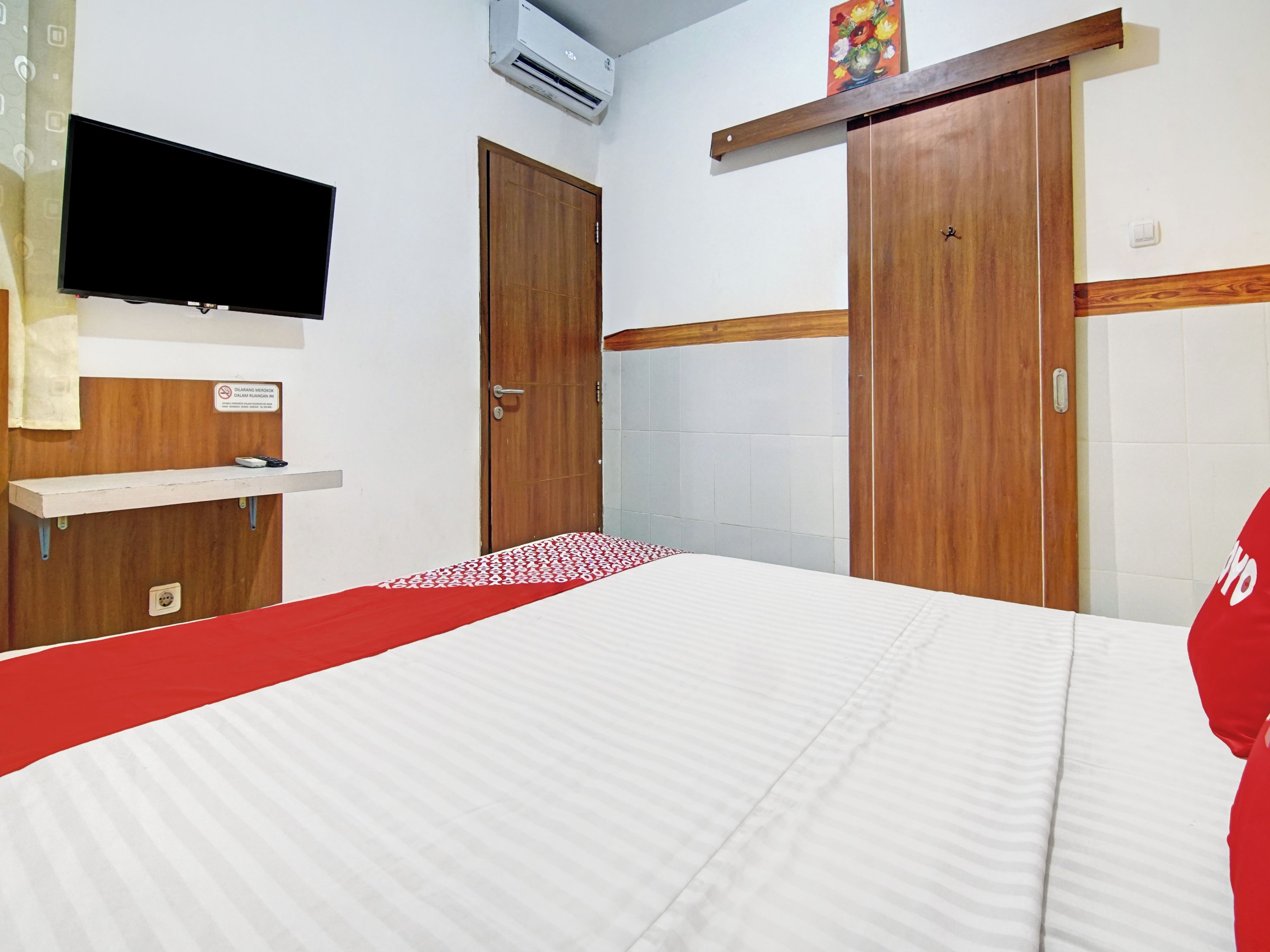 Hotellihuone, jossa on sänky, seinälle asennettu televisio, puinen vaatekaappi ja hylly, jonka päällä on taulu.